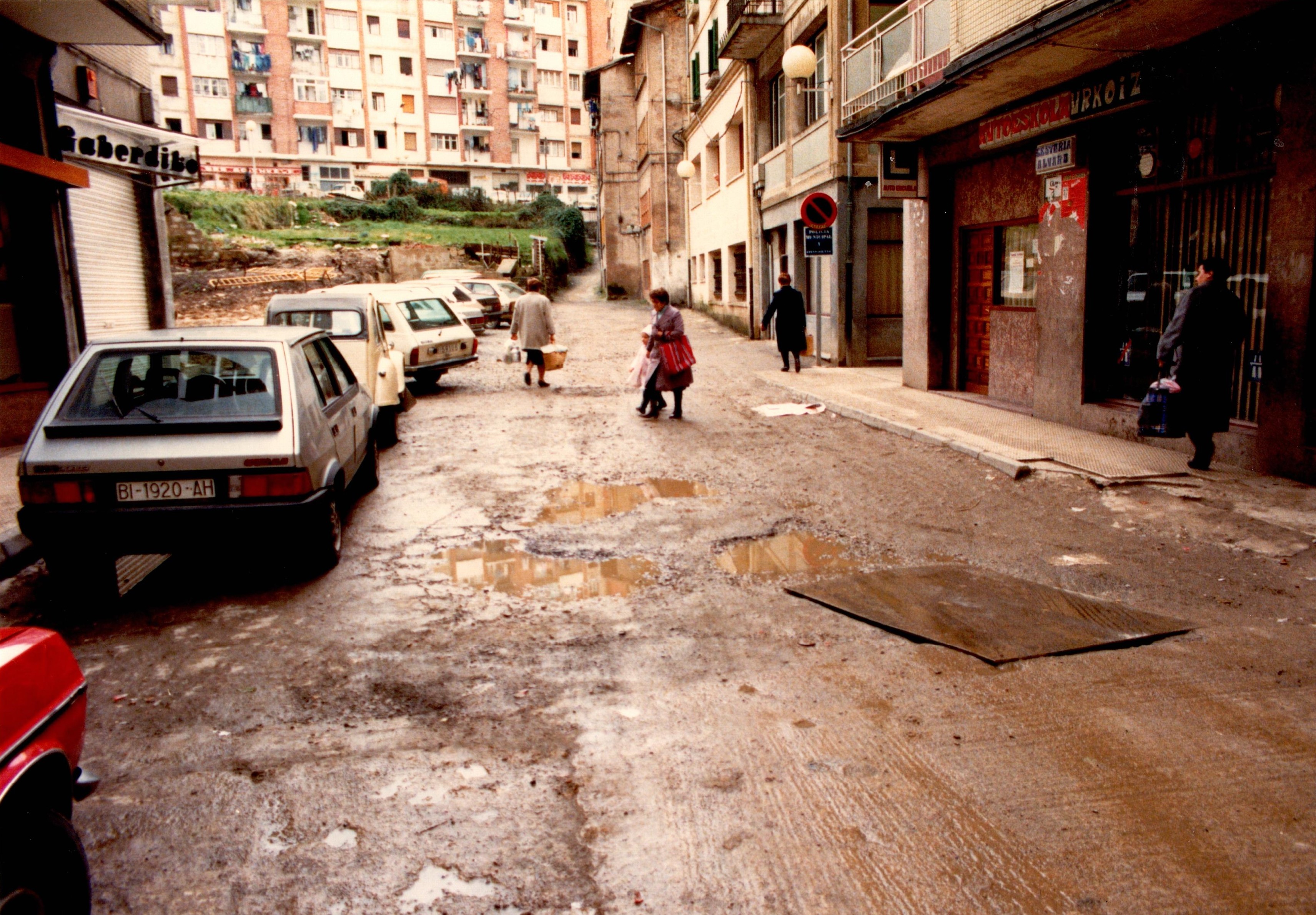 Se ve una calle con el asfalto en malas condiciones, llenas de charcos y baches. A la izquierda hay coches aparcados y por mitad de la calle y en las aceras gente transitando. A la izquierda, después de unos edificios en una parcela hay vallas amarillas y las obras ya han comenzado porque se ve parte de las herramientas.