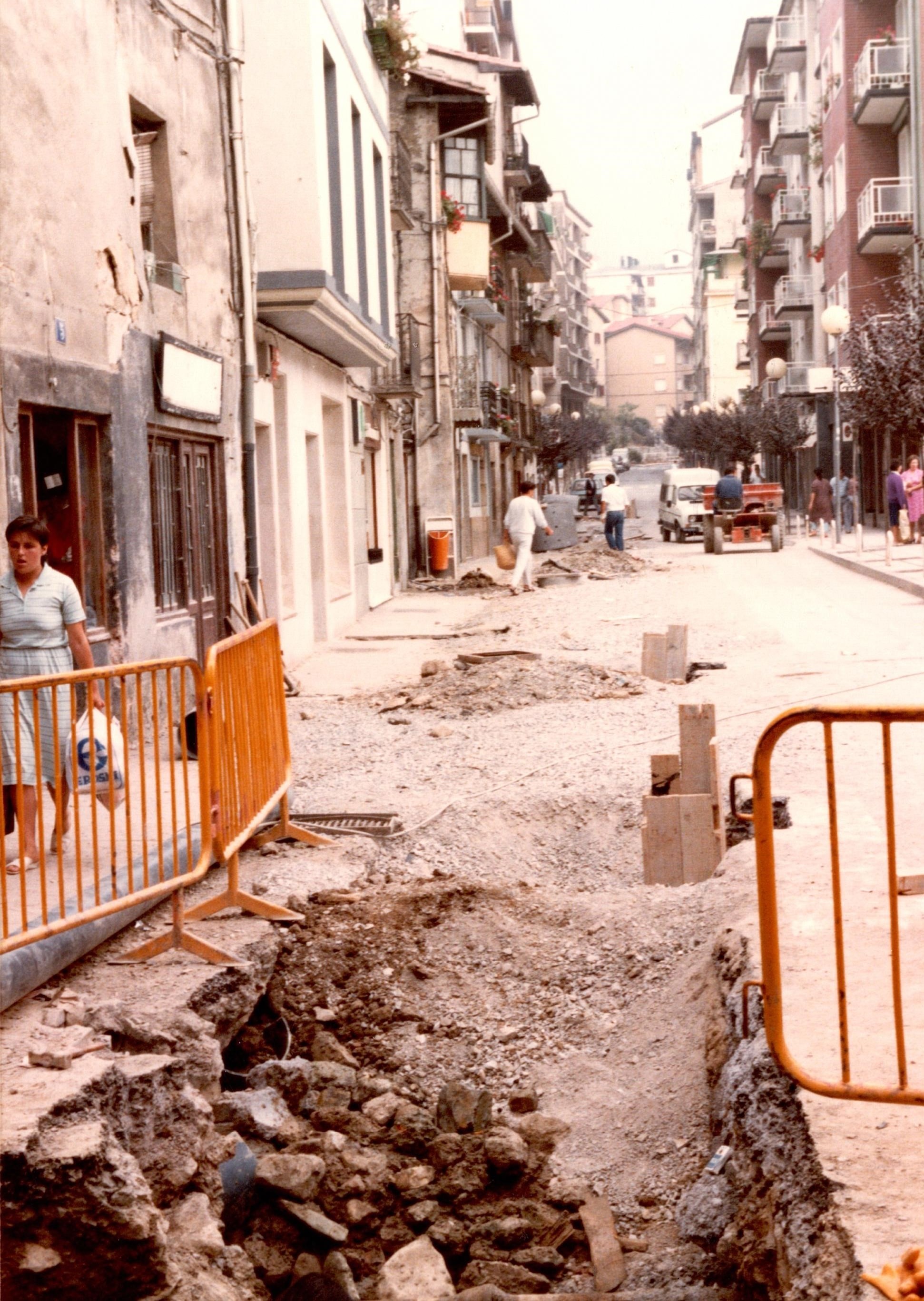 Se ve el asfalto de la calle Zubiaurre, la carretera levantada con escombros y vallas amarillas. A la izquierda una señora camina con bolsas en la mano por la acera y al fondo puede verse maquinaria de la obra y algunos obreros.
