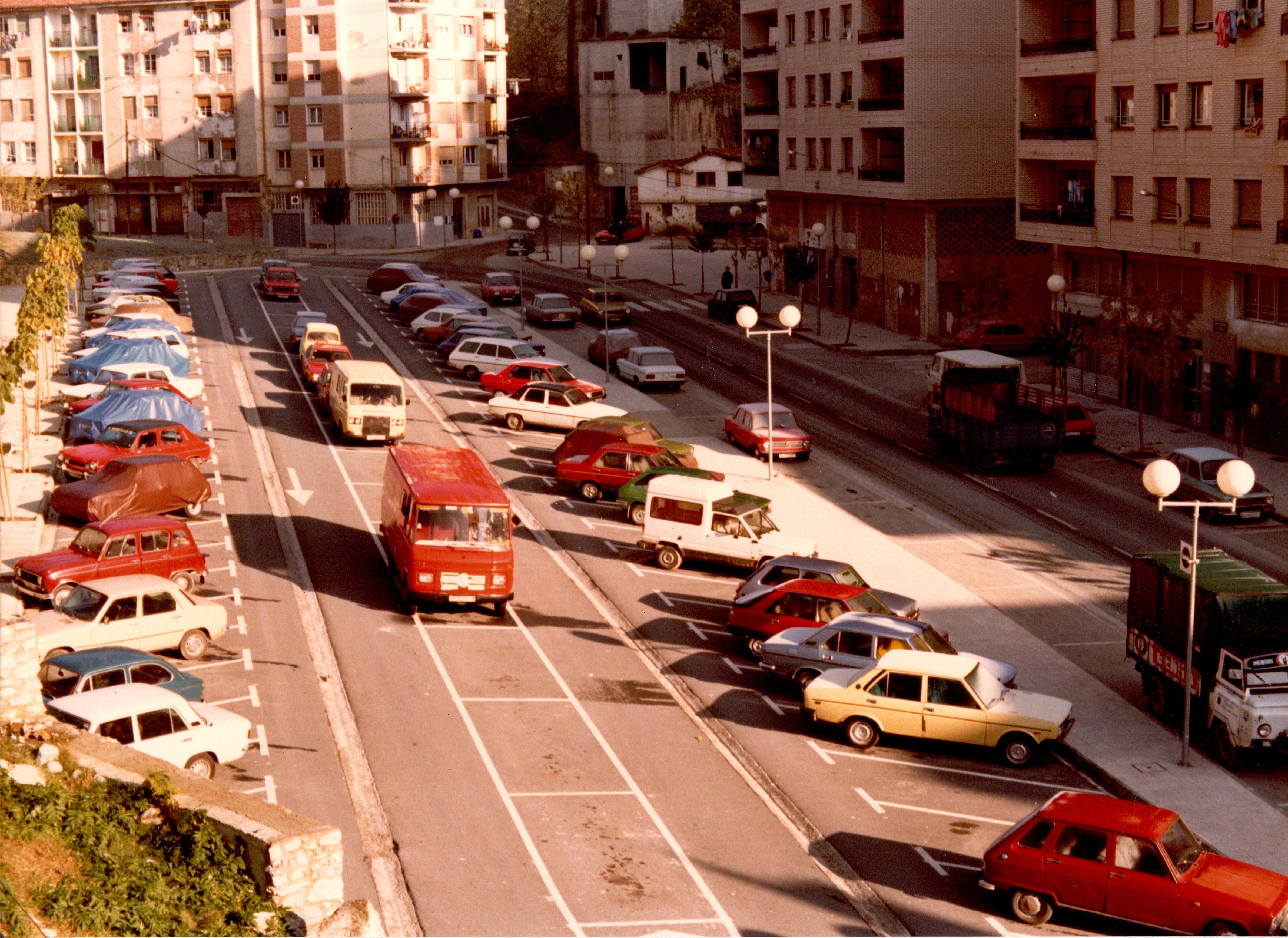 Se ve un aparcamiento con estacionamientos en batería y paralelo. Hay coches de muchos colores: crema, blanco, verde, azul, rojo, negro... también hay alguna furgoneta y camión. Al fondo y en la zona derecha hay edificios, viviendas.