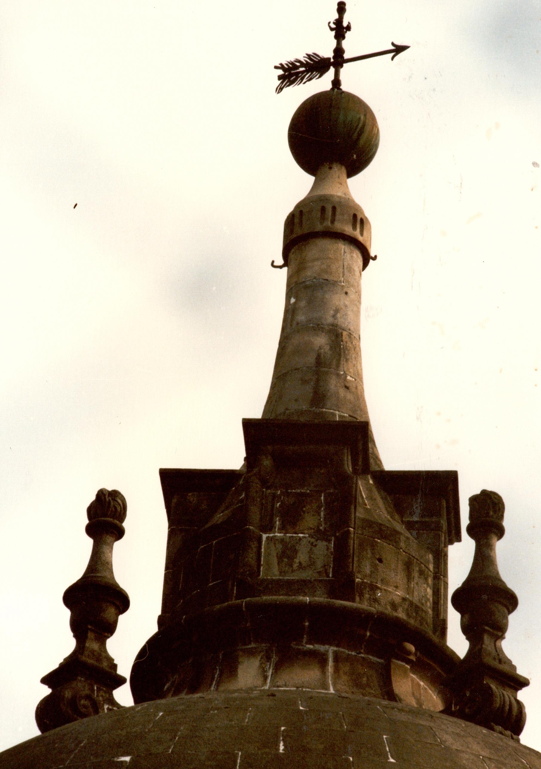 Se muestra la parte superior de la cúpula del palacio, es una fotografía hecha desde el tejado, de cerca. Se ven dos pináculos, la esfera superior y la veleta como elemento más alto decorativo. Es una estructura de piedra, de color marrón oscuro y claro según donde le de la luz. En el fondo se ve el cielo.