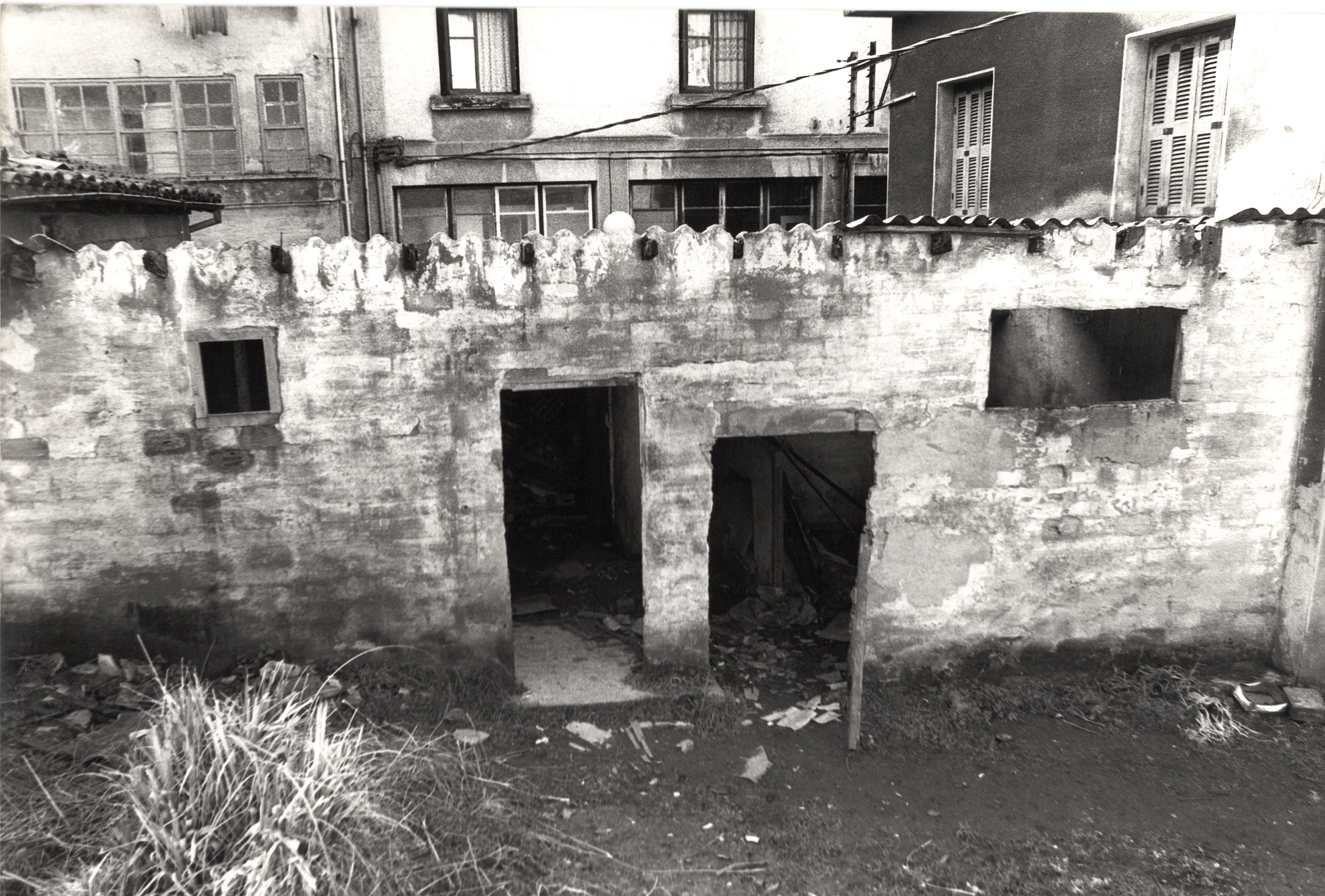 Se ve una fachada bastante deteriorada de una estructura/edificio de piedra, hay dos puertas, una más alta que la otra pero sin marco ni puerta de acceso, es solo la forma. También hay dos ventanas. Al fondo se ven más edificios.