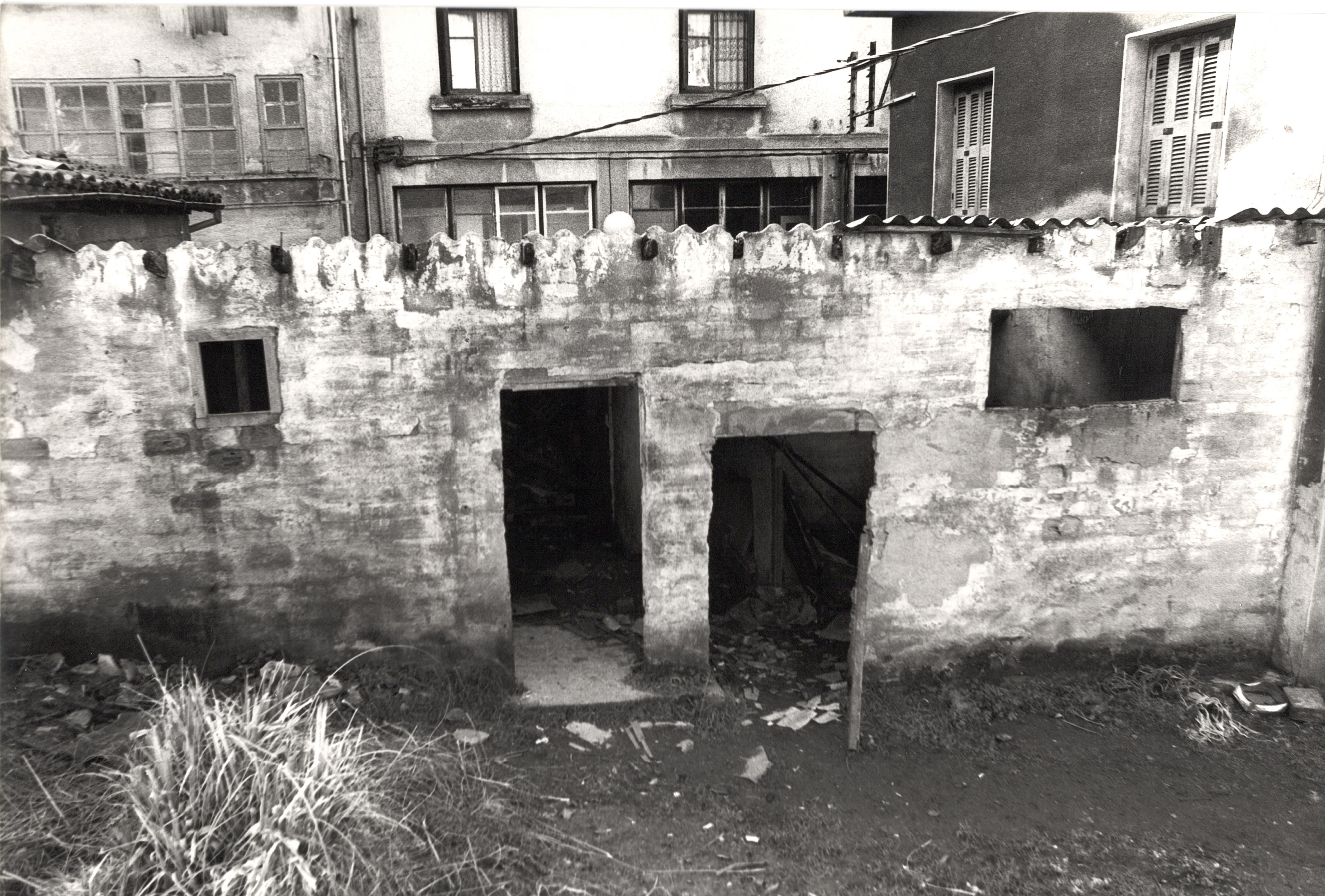 Se ve una fachada bastante deteriorada de una estructura/edificio de piedra, hay dos puertas, una más alta que la otra pero sin marco ni puerta de acceso, es solo la forma. También hay dos ventanas. Al fondo se ven más edificios. 