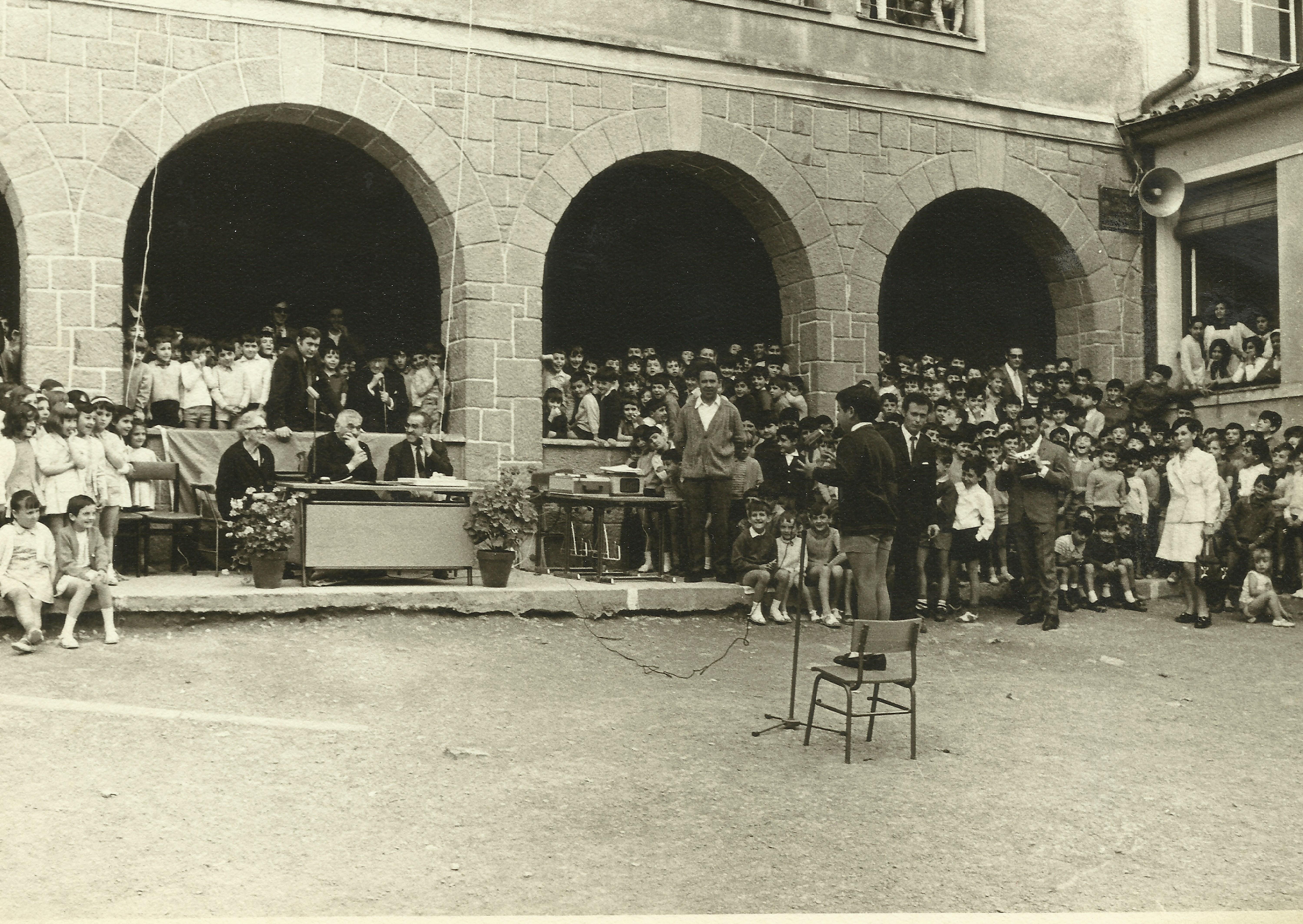 Fotografía en blanco y negro del Colegio Teresa Murga. Hay un niño frente a un micrófono y público en pie y sentados en la zona de los soportales y ventanas. 