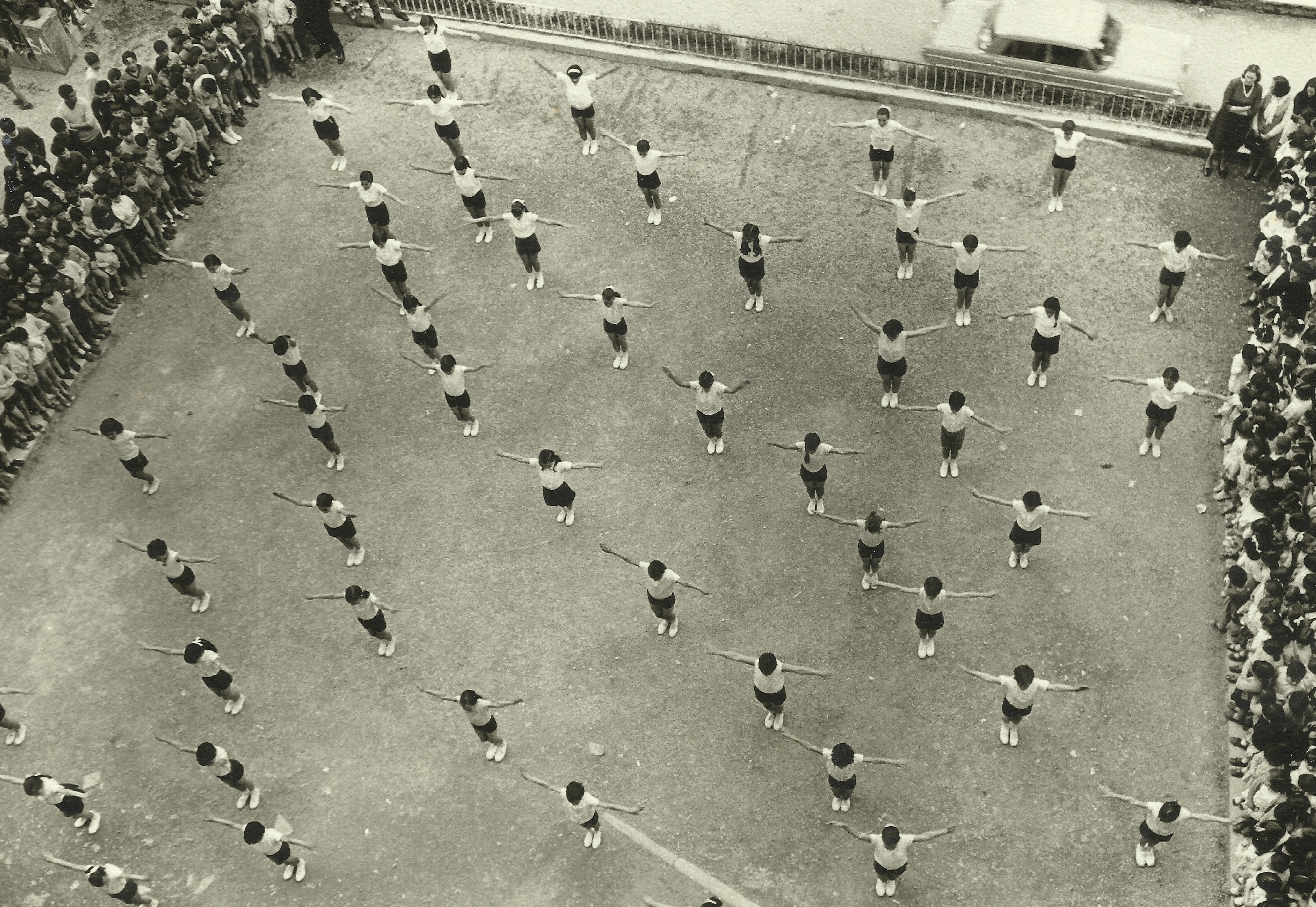 Fotografía en blanco y negro con ángulo cenital de unas 50 chicas aproximadamente haciendo un espectáculo de gimnasia en el patio del Colegio Teresa Murga. A lados hay pública viendo el ejercicio que hacen las chicas.