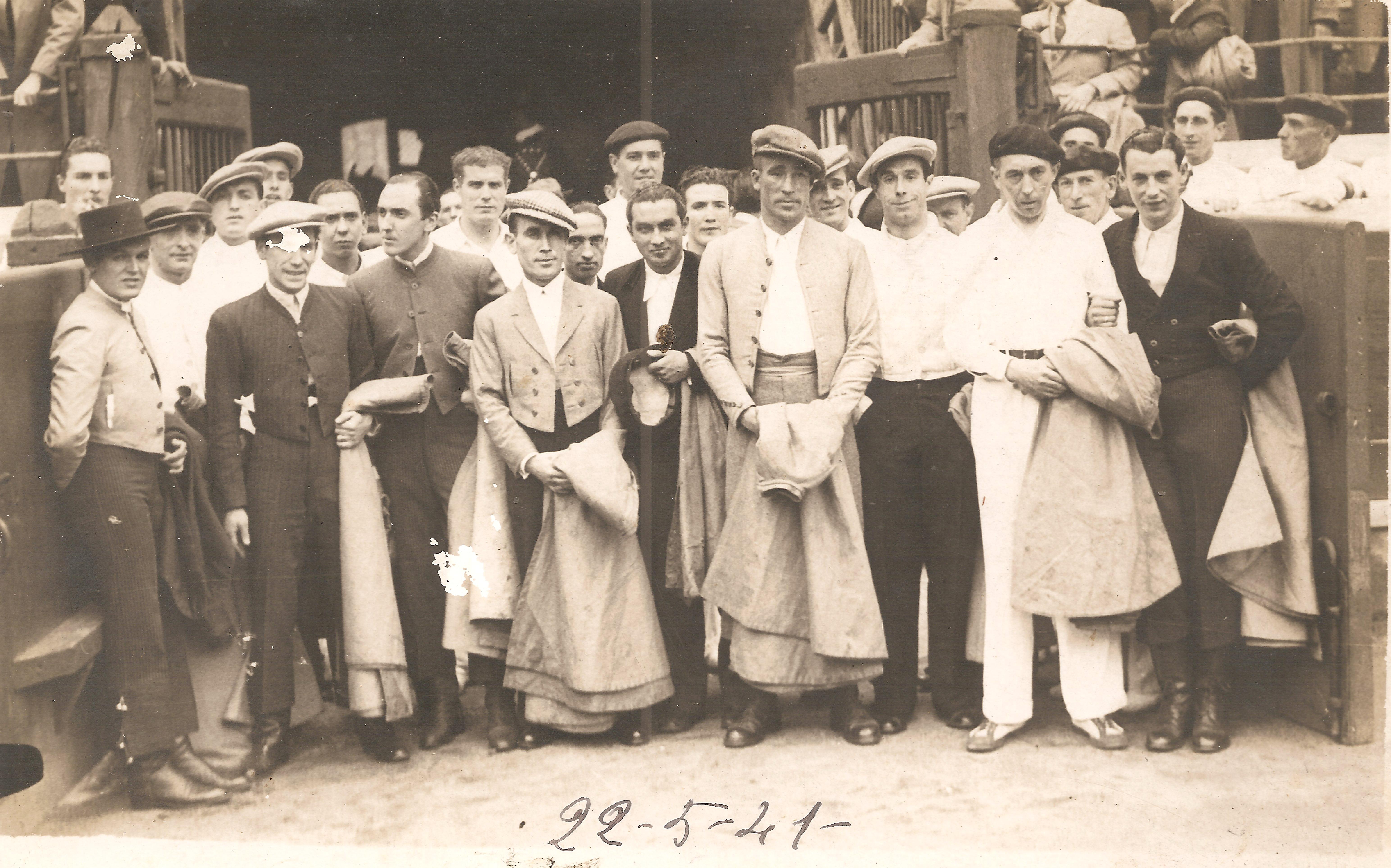 Félix Izagirre junto con otros 25 hombres, algunos visten con el traje típico de torero y capote en mano, otros llevan txapela y otros sombrero. Félix y otros visten de blanco tanto camisa como pantalón. Fotografía en blanco y negro