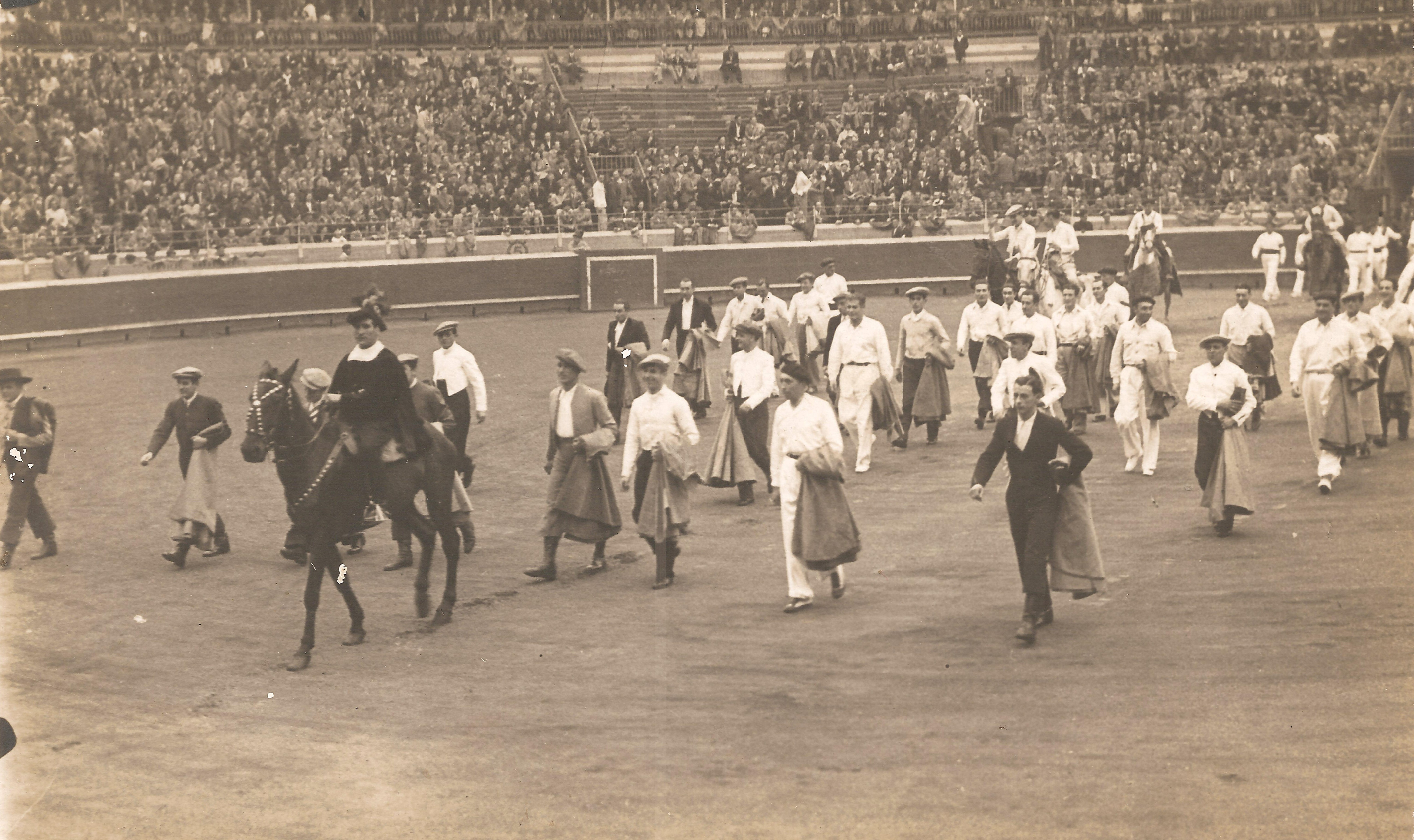 Fotografía en blanco y negro donde están caminando por el ruedo de la plaza de toros. Es un grupo grande de hombres entre los que se encuentran otros pelotaris como Félix Izagirre, toreros y cinco van a caballo, a diferencia del resto que va a pie. En el fondo se ven el burladero, la barrera y el tendido donde está el público.