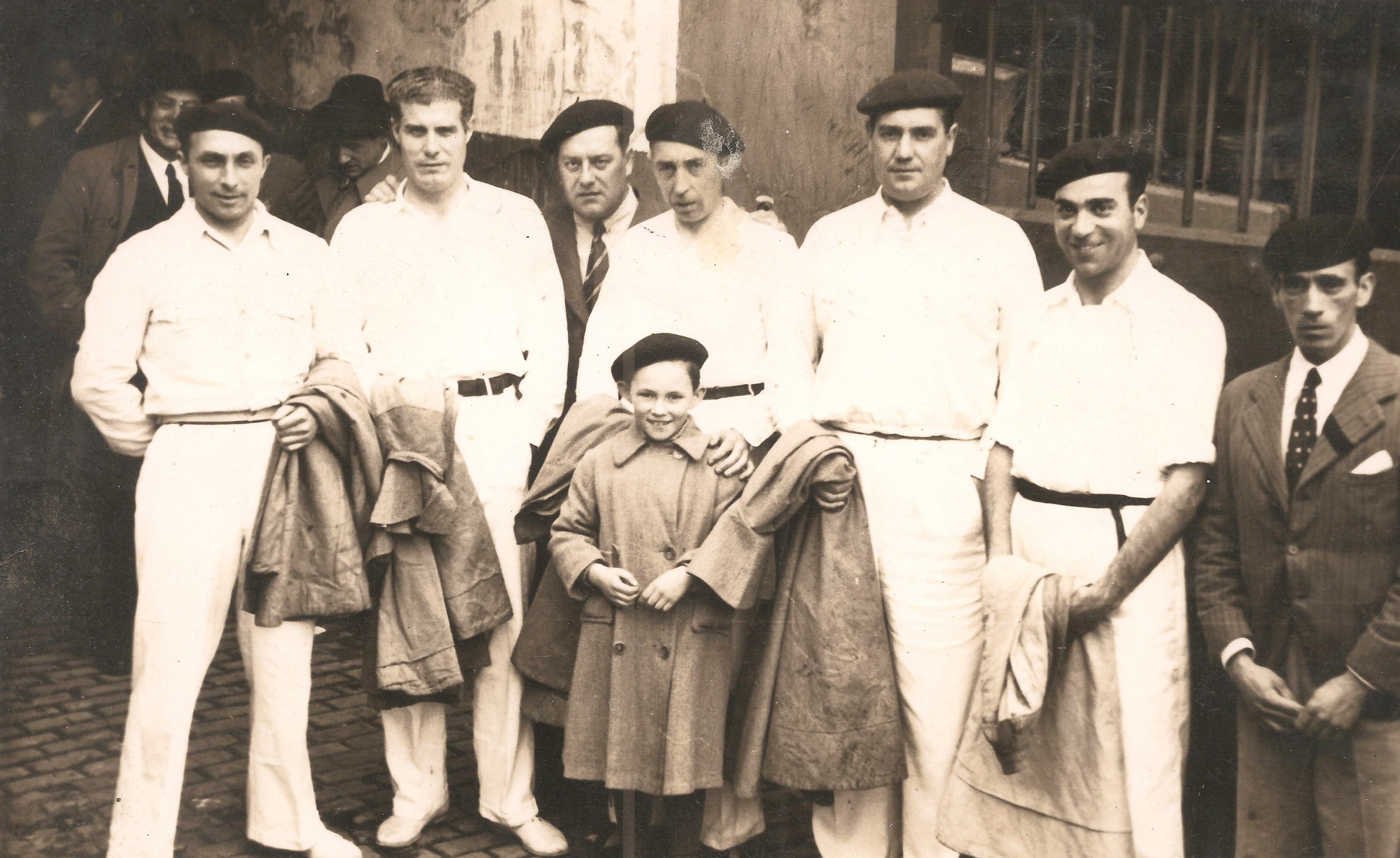 Félix Izagirre acompañado de otros hombres el día que estuvo en la plaza de toros de Bilbao. Hay siete hombres y un niño, cinco visten de blanco, dos hombres de traje y corbata. Todos llevan txapela menos Félix Izaguirre Garate. La fotografía es en blanco y negro.