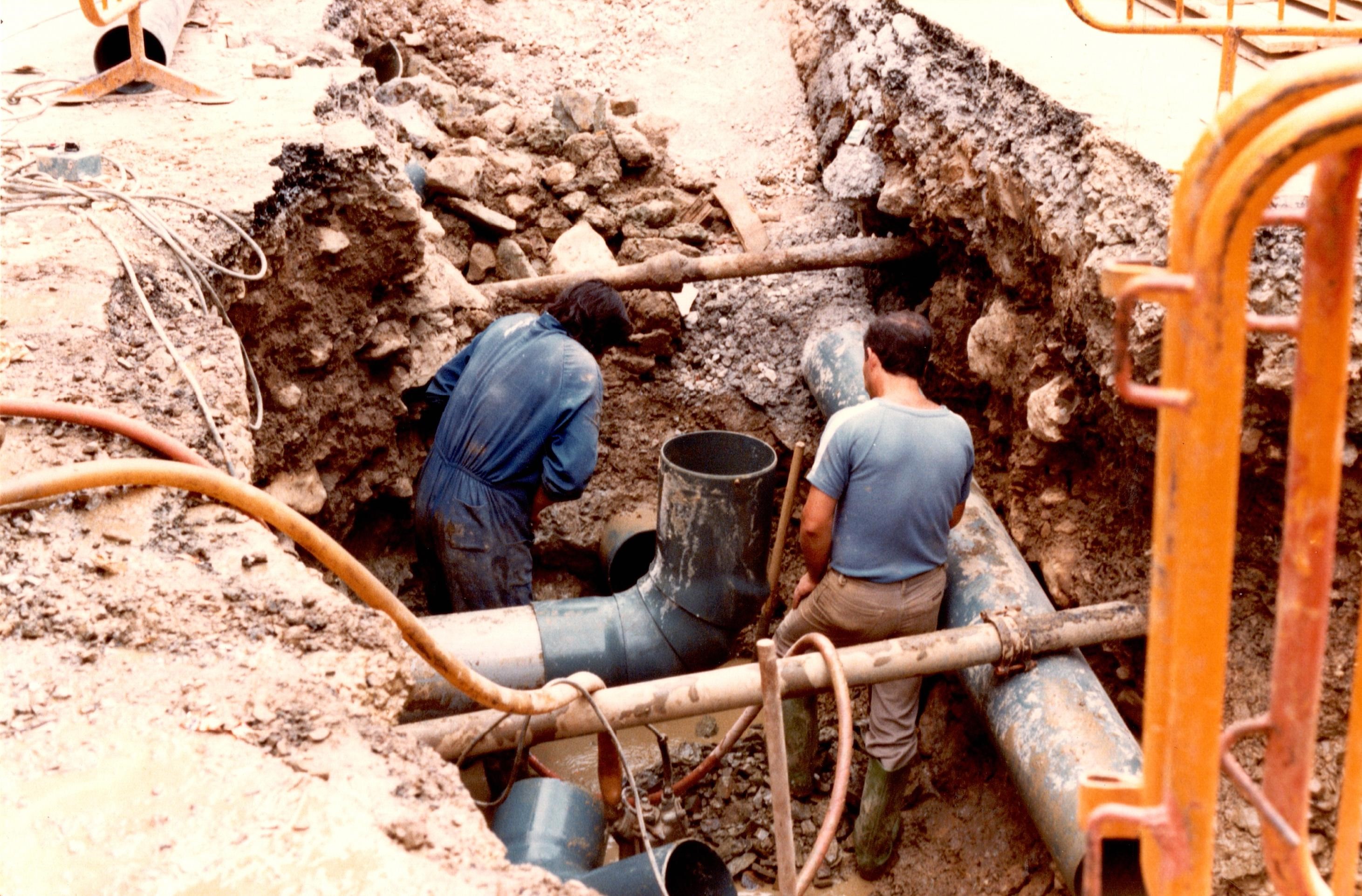 Dos obreros están dentro de una zanja trabajando en la obra de las tuberías. Uno de ellos lleva un mono azul de obra yel otro botas, pantalón marrón/gris y camiseta de manga corta azul. Se ven algunas tuberías y escombro. También hay algunas vallas amarillas encima, en la acera y no dentro de la zanja. 