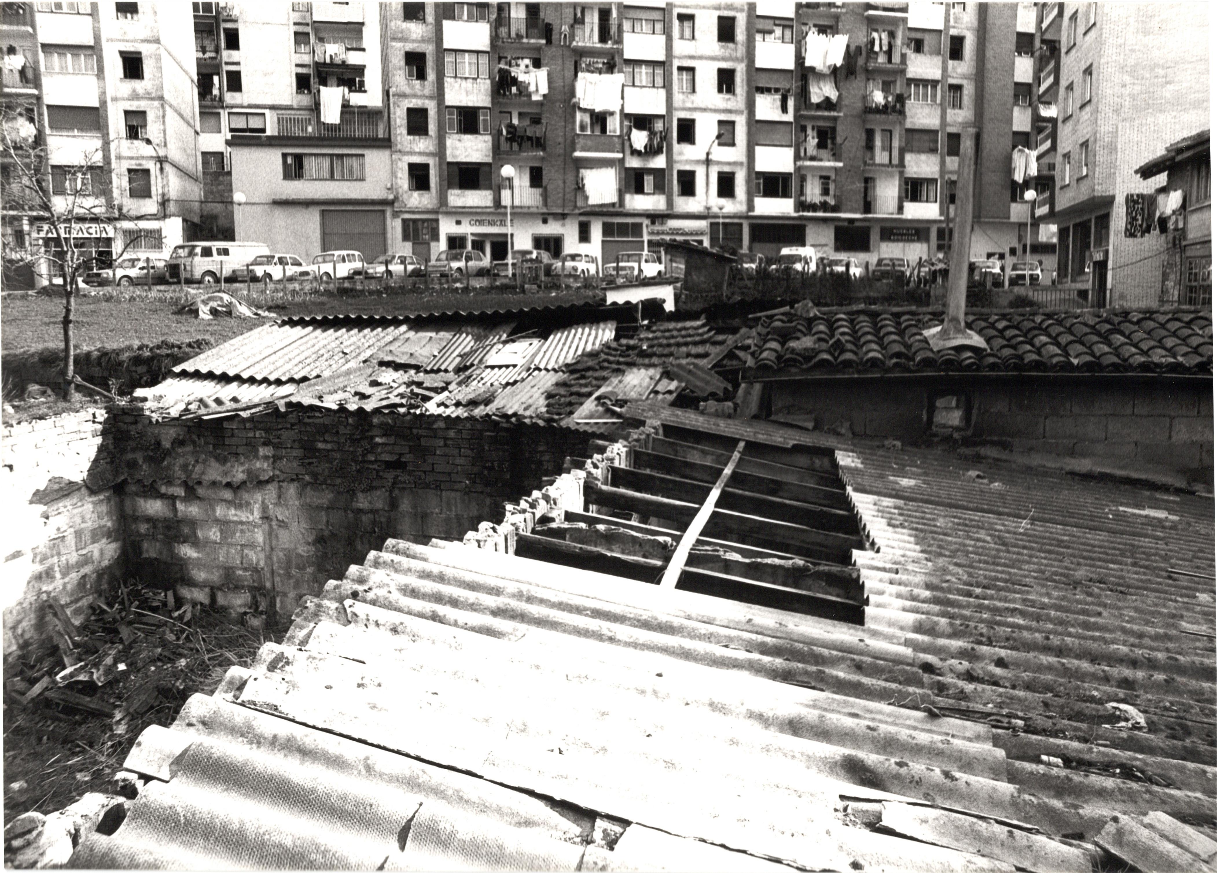 Se ven los edificios de unas chabolas/casetas hechas de madera y piedra, algunos tejados son de teja pero tros tienen plásticos que asemejan la teja. En uno de esos tejados hay una chimenea, y otro tiene agujeros. Al fondo se ven los edificios de la calle Goienkale y los bajos de los mismos. Se puede leer "farmacia" en uno de los locales, en otro "Goienkale". La fotografía es en blanco y negro. En la calle del fondo, la calle Goienkale, hay varios coches aparcados en batería.
