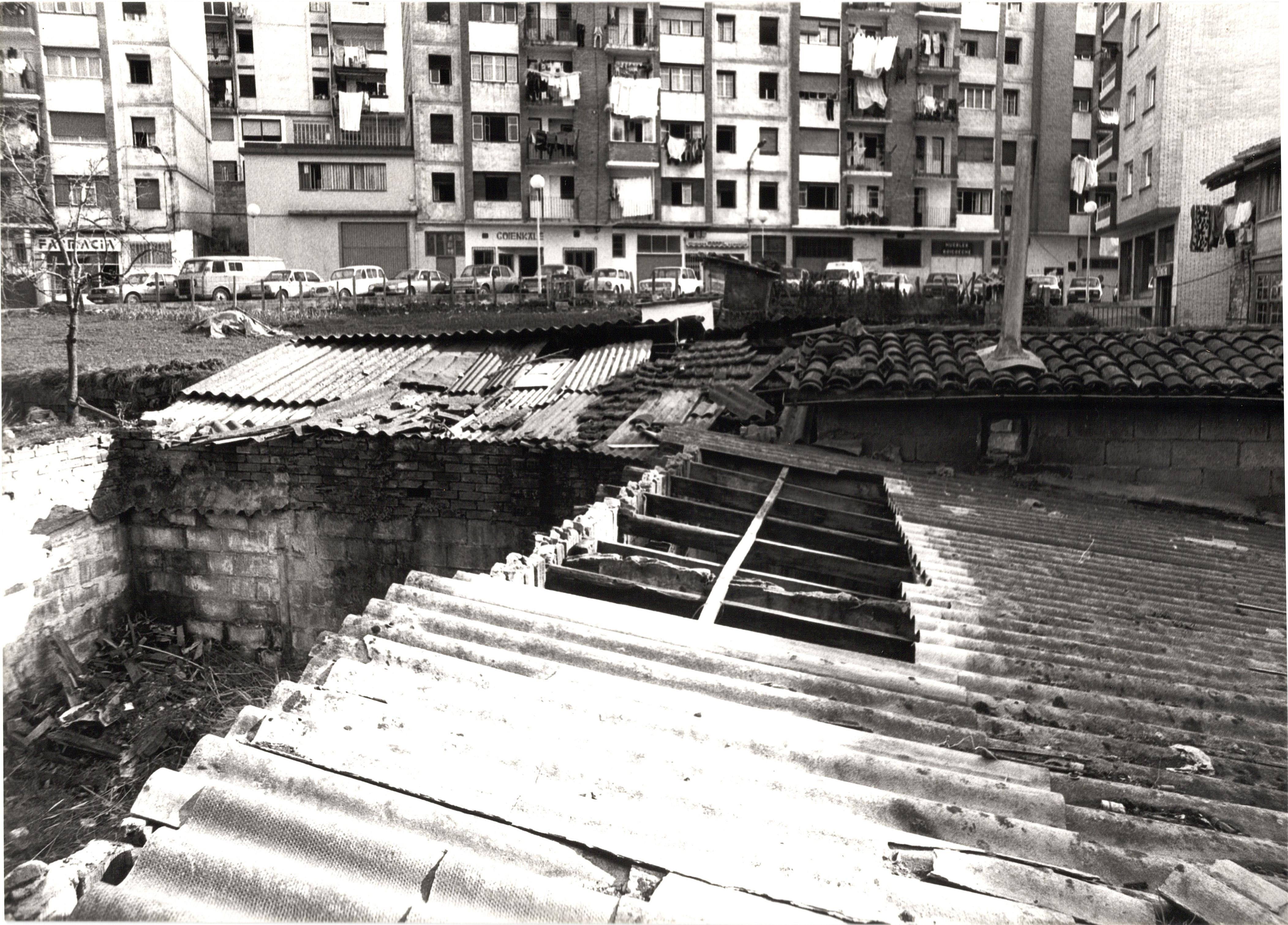 Se ven los edificios de unas chabolas/casetas hechas de madera y piedra, algunos tejados son de teja pero tros tienen plásticos que asemejan la teja. En uno de esos tejados hay una chimenea, y otro tiene agujeros.  Al fondo se ven los edificios de la calle Goienkale y los bajos de los mismos. Se puede leer "farmacia" en uno de los locales, en otro "Goienkale". La fotografía es en blanco y negro. En la calle del fondo, la calle Goienkale, hay varios coches aparcados en batería. 