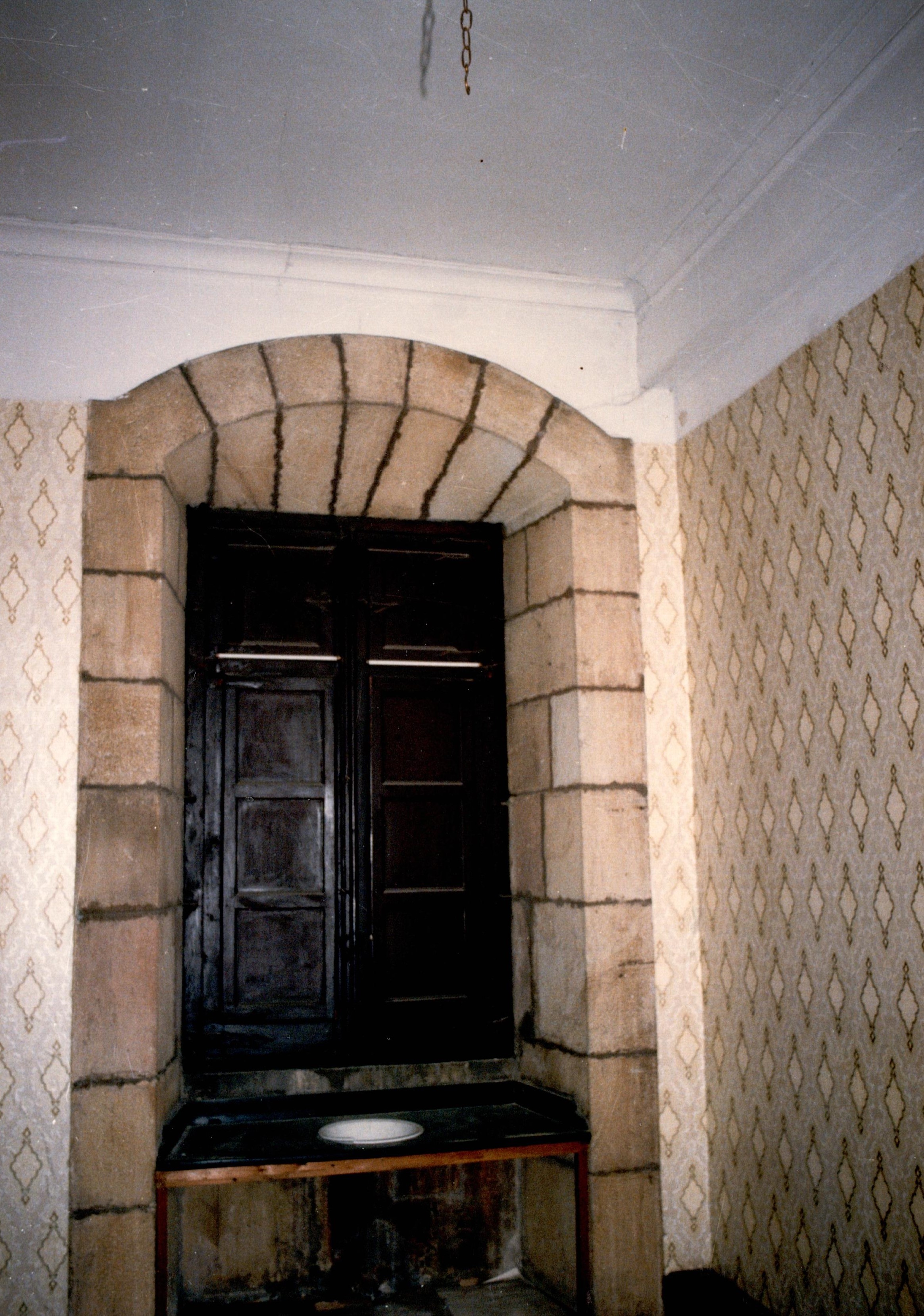 Se muestran las contraventanas de una ventana, son de madera y de color oscuro. A los lados se ven las paredes con lo que parece papel con decoraciones en forma de rombo pero con el borde irregular. Ese papel es amarillo suave y marrón.