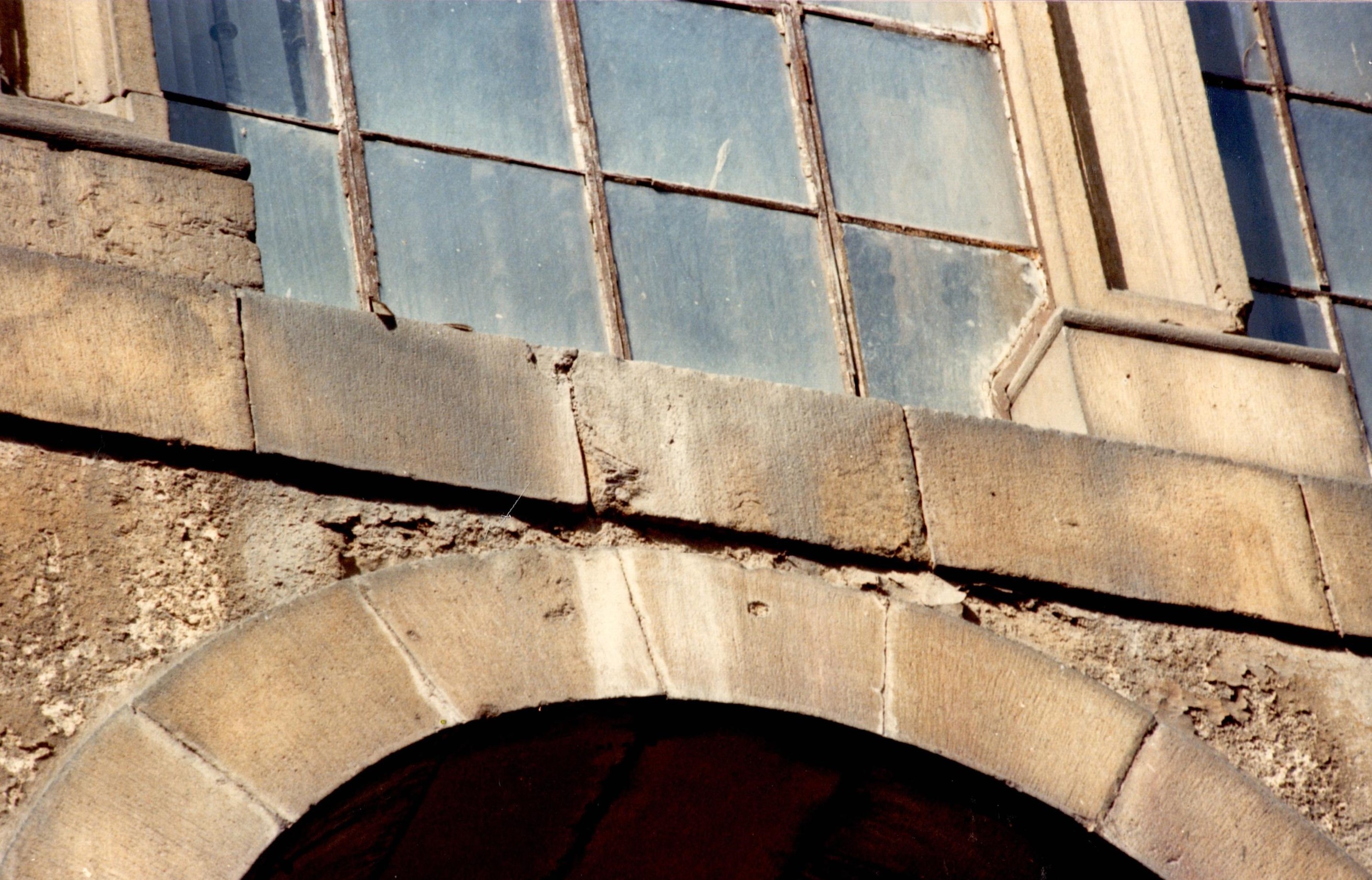 Se ve parte de un arco, solo la parte superior del exterior del palacio y unas ventanas en condiciones no muy buenas, sucias, con una capa blanca de polvo y suciedad. Se ve el color y el material del edificio, un color arena tostada, clarita.