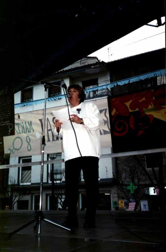 María Rosario Arrizabalga Urreta está sobre una escenario dando un discurso frente a un micrófono, tiene un papel entre sus manos , viste con un jersey blanco y pantalones negros. 