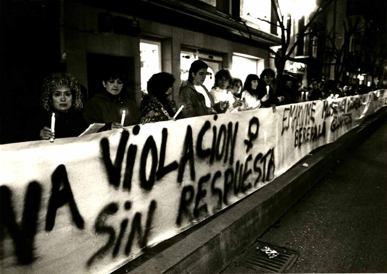 Varias mujeres están tras una pancarta en la que se puede leer: una violación sin respuestas y más texto en euskera que no se puede leer del todo (palabras como emakume y berehala). Cada una de ellas tiene una vela en la mano encendida. Están encima de una acera y delante de ellas está la carretera.