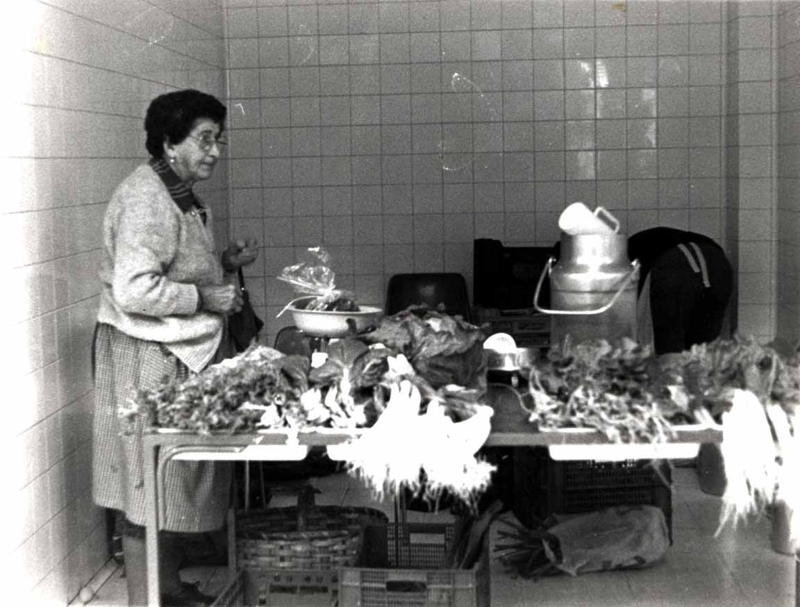 Fotografía en blanco y negro donde se ve a una señora a la izquierda de pie. La señora lleva falda hasta la rodilla o un poco más abajo incluso, una chaqueta y por debajo una camisa que puede apreciarse mirándole el cuello. Tiene pendientes, pelo oscuro y gafas. Se ve un pequeño puesto a su derecha con hortalizas y otras verduras junto con un peso para la venta de la materia. El fondo es una pared de azulejos (aparentemente blanca o gris). En el suelo, debajo de la mesa, hay cestas de madera y plástico.
