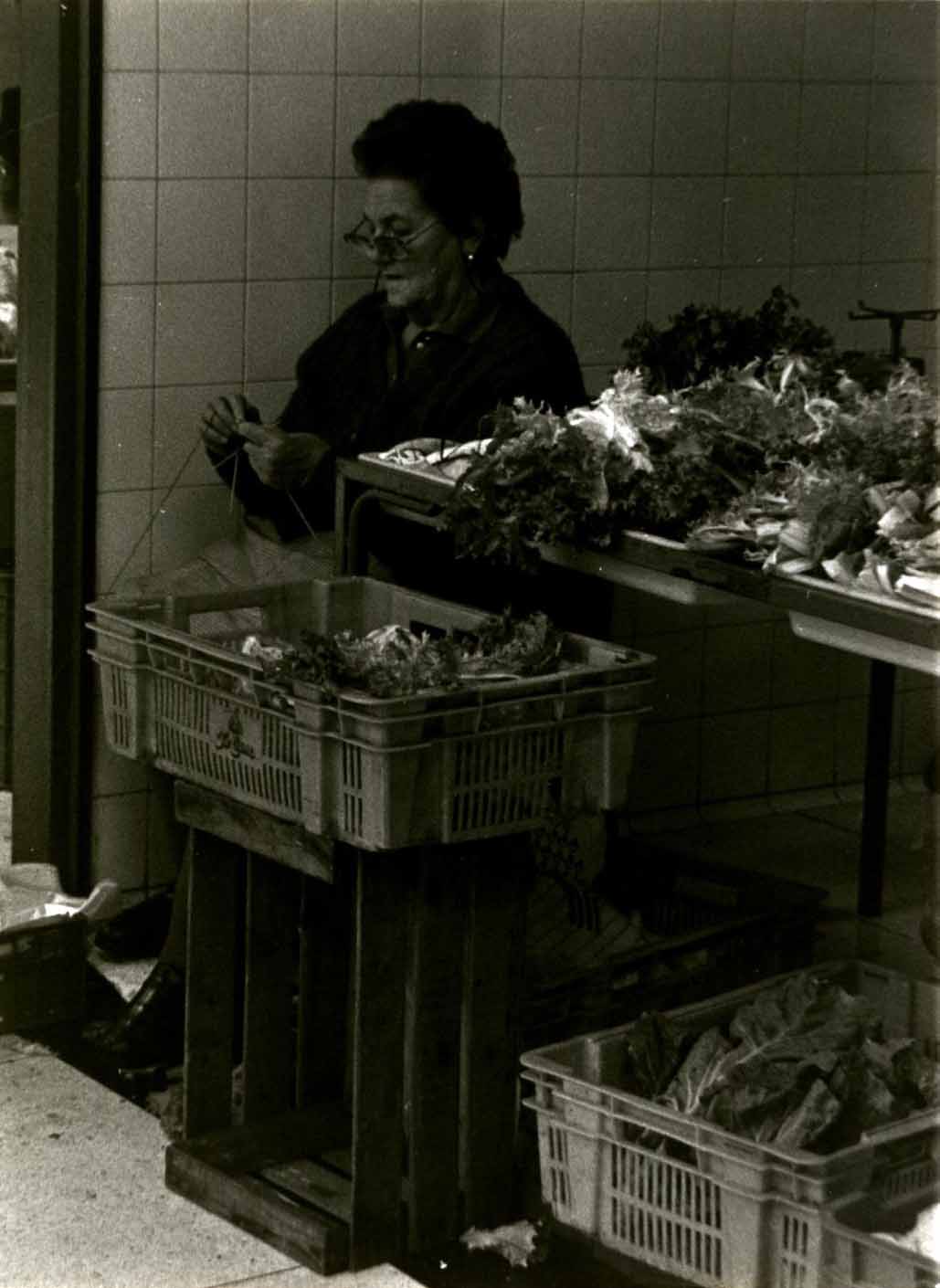 Una mujer está sentada a la izquierda de su puesto, tejiendo o pasando el rato con alguna manualidad. A su derecha está el puesto donde pueden vrse lechugas, acelgas y otras verduras y hortalizas. La fotografía es en blanco y negro. La señora tiene pelo oscuro, gafas y pendientes. 