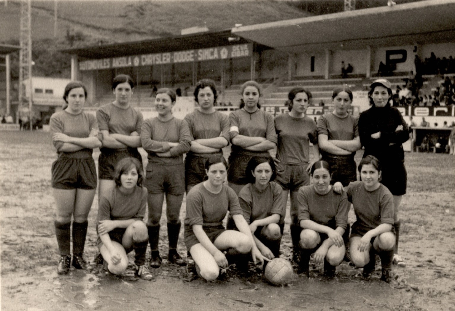 Equipo de fútbol femenino en el campo, sobre el barro mojado. Son trece mujeres en total y de fondo se ven las gradas del campo