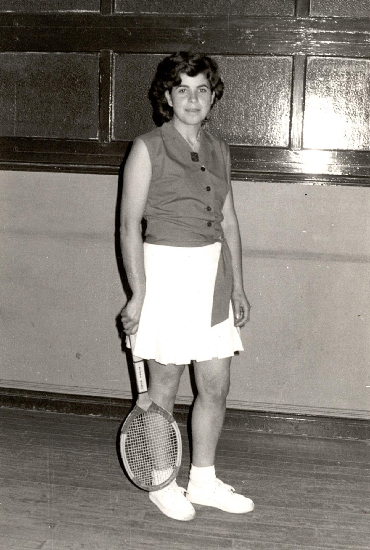 Fotografía en blanco y negro de la raquetista Chari. Viste con falda blanca, zapatillas deportivas blancas y camisa de manga corta. En su mano derecha sujeta la raqueta desde el mango. 