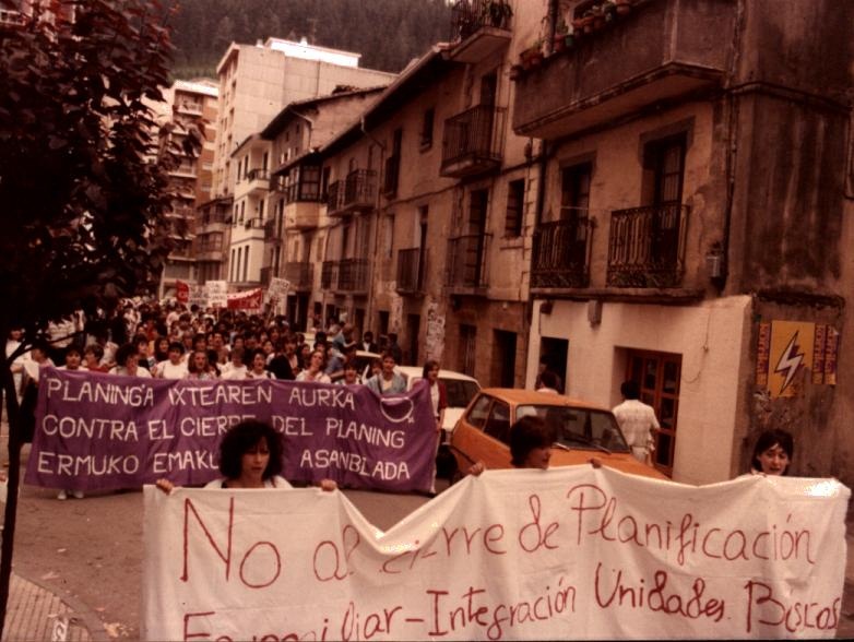Un grupo de personas van caminando por la calle Zubiaurre. Se ven algunos coches y los edificios a la derecha. Hay una primera pancarta sujeta por tres mujeres donde se puede leer "no al cierre de Planificación Familiar-Integración Unidades" de fondo blanco y letras rojas. Detrás va otra pancarta de fondo morado y letras blancas donde puede leerse "Planing-a ixtearen aurka, contra el cierre del planing, ermuko emakume asanblada". 