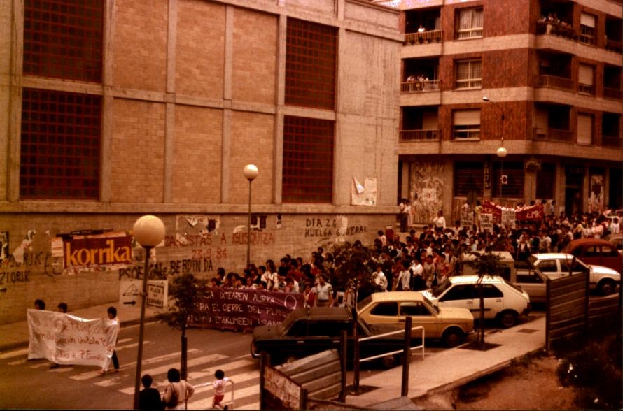 Un grupo de personas van caminando por la calle. Hay dos pancartas, la primera tiene el fondo blanco con letras rojas. La segunda es morada con letras blancas. Tras la primera pancarta van tres personas y tras la morada el resto de personas. Hay coches aparcados, se ve el edificio del mercado y algunas pancartas y pintadas en la fachada del mercado.  