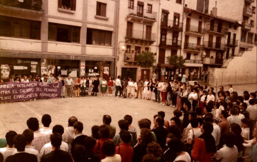 Un grupo amplio de personas forman un círculo en la plaza Cardenal Orbe. Hay una pancarta donde se puede leer "Planing"a ixtearen aurka. Contra el cierre del planing. Ermuko Emakumeen Asanblada. Hay hombres y mujeres en el círculo, la foto es a color y hay aproximadamente más de 100 personas
