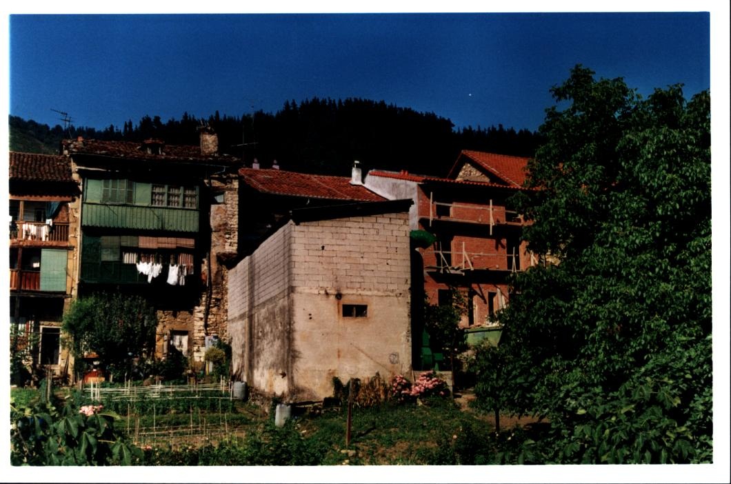 Fotografía de la parte trasera de la Casa de la Mujer, se ve una fachada ampliada, sin terminar y a su izquierda y derecha huertas, árboles y otras casas o edificios. La foto es a color. 