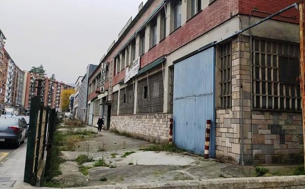 Se ve un edificio envejecido con un gran cartel con fondo blanco y letras negras en el que pone "Se vende" y un número de teléfono. También hay un cartel en vertical donde se puede leer "Idesa". Delante del edificio hay asfalto con grietas, algunos espacios con hierba que brota del asfalto.  Hay una verja que separa el terreno del edificio con una acera y una carretera, donde hay algunos coches aparcados. A la izquierda se aprecian varios bloques de viviendas. 